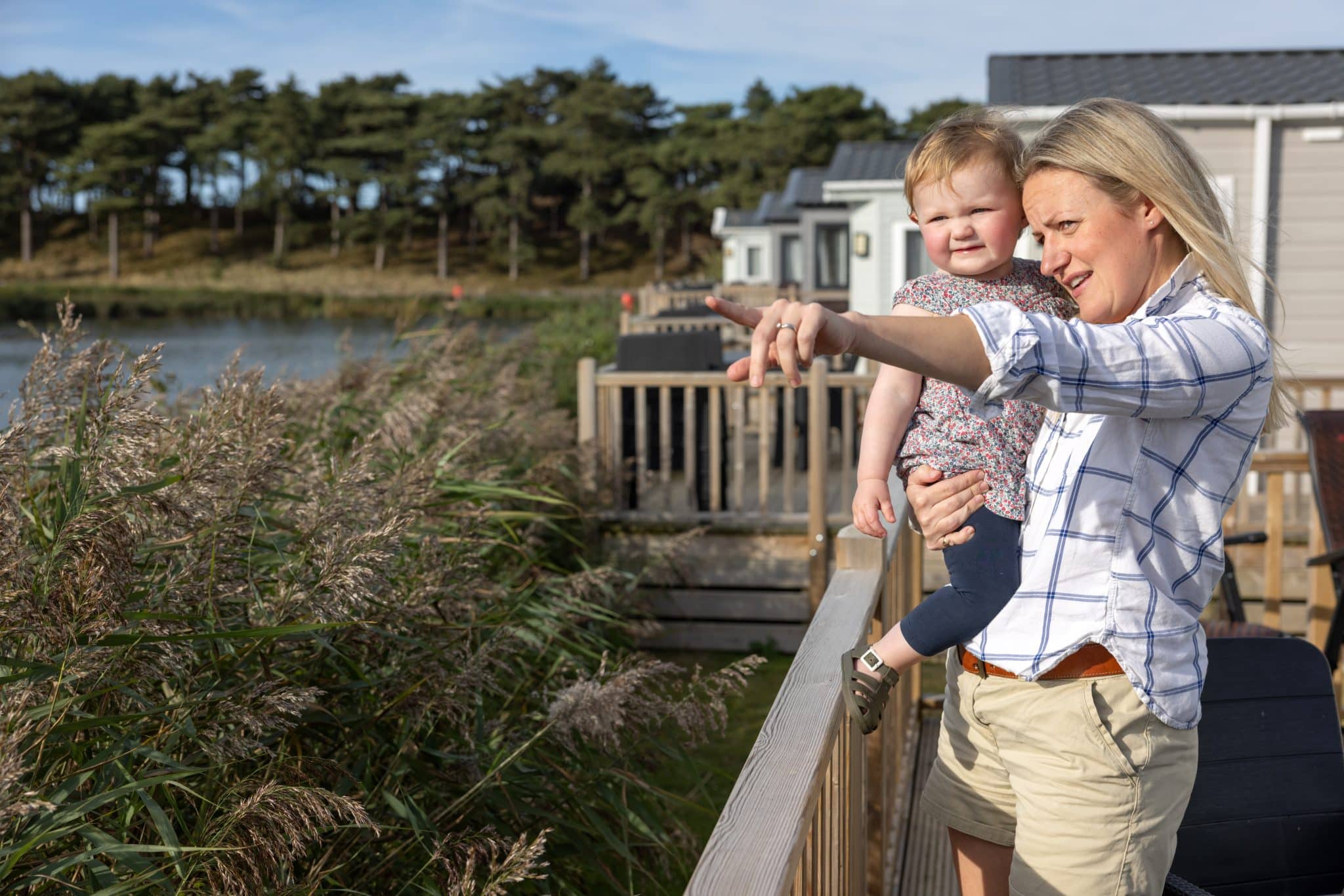 A mum holds her baby and points out towards a lake from the decking of a Pinewoods caravan.