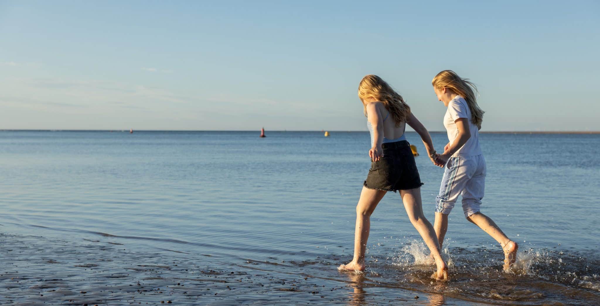 Two girls in summer clothes run in the shallows of the sea.