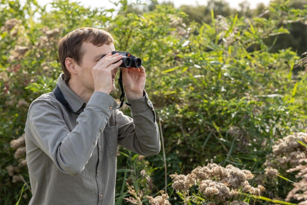 A man in a green shirt stands looking through binoculars.