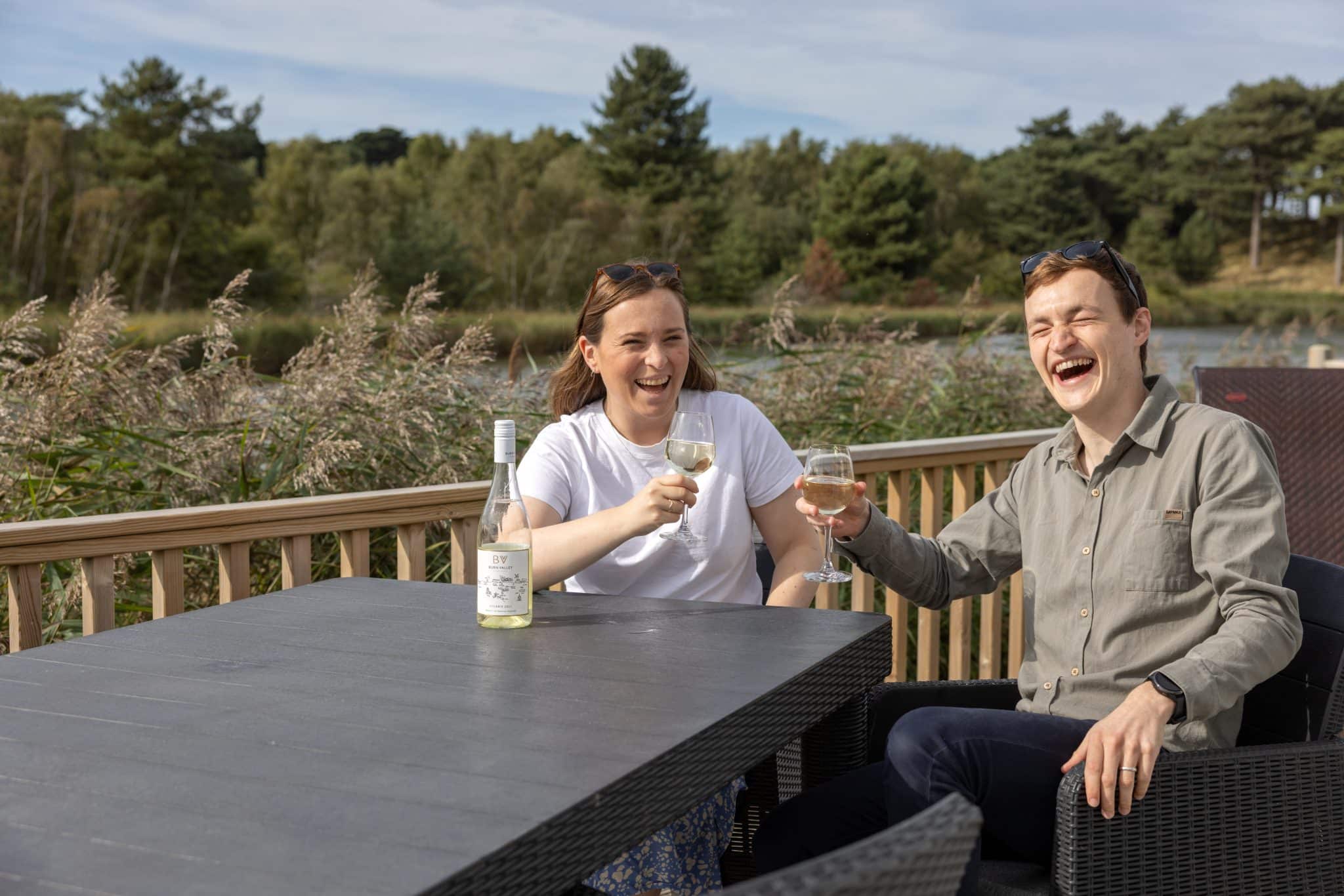 A couple sit at a table on the decking with a glass of white wine each, laughing.