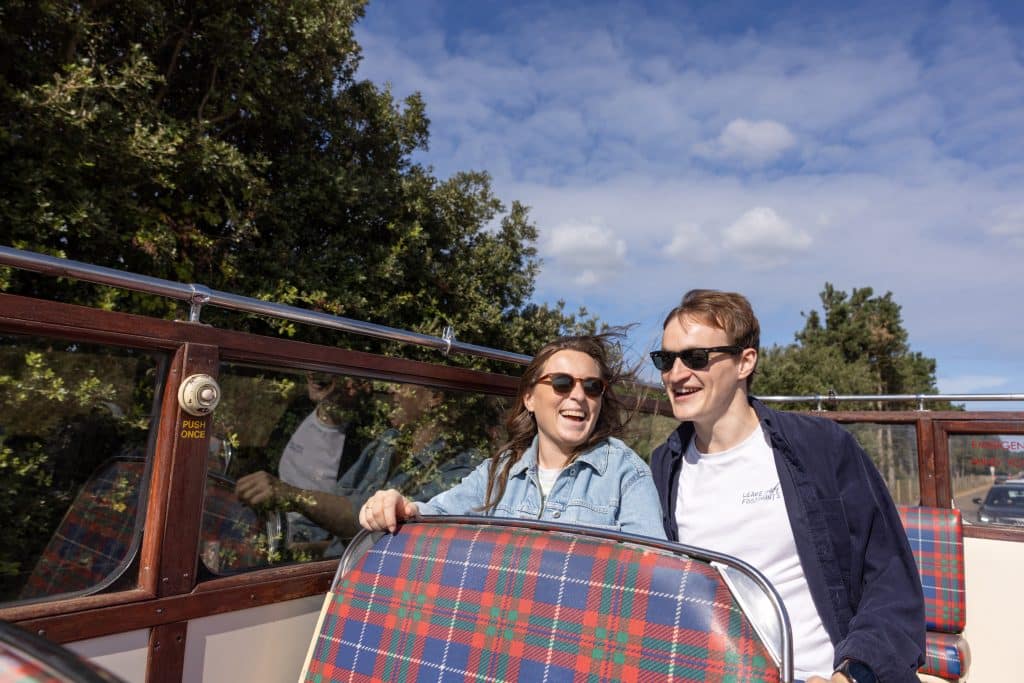 A couple in their early thirties in sunglasses laugh on a vintage, open-topped bus.