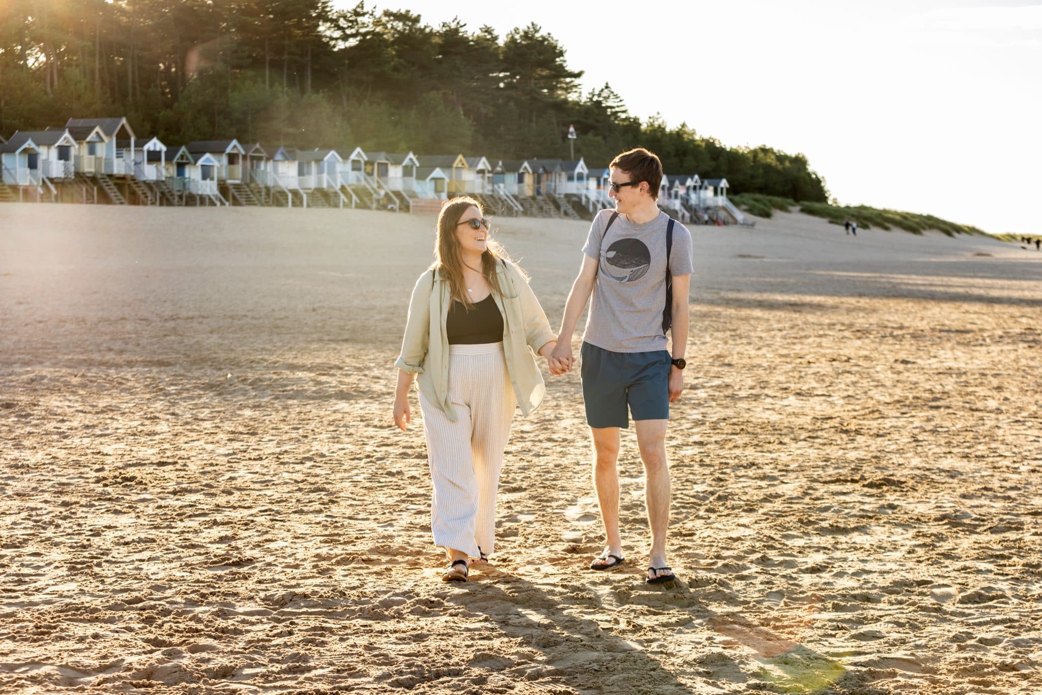 A couple walk along sandy Wells Beach, with the beach huts in the background.