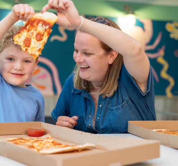 A woman and a little boy hold up a slice of pepperoni pizza, smiling.