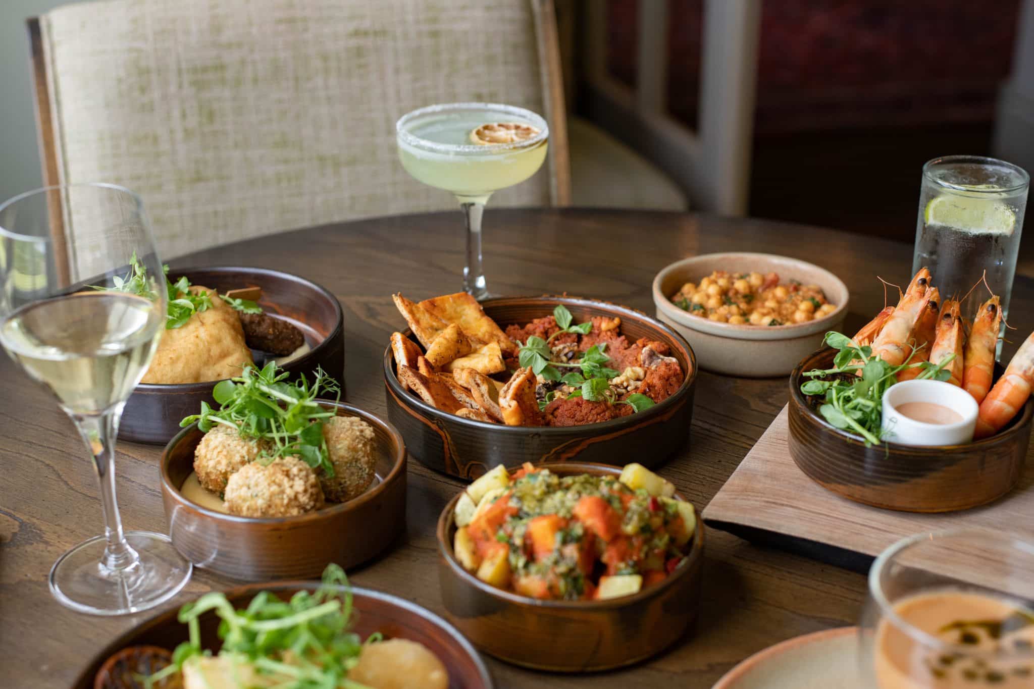 A selection of small, tapas style dishes on a wooden table alongside a cocktail and glass of wine.