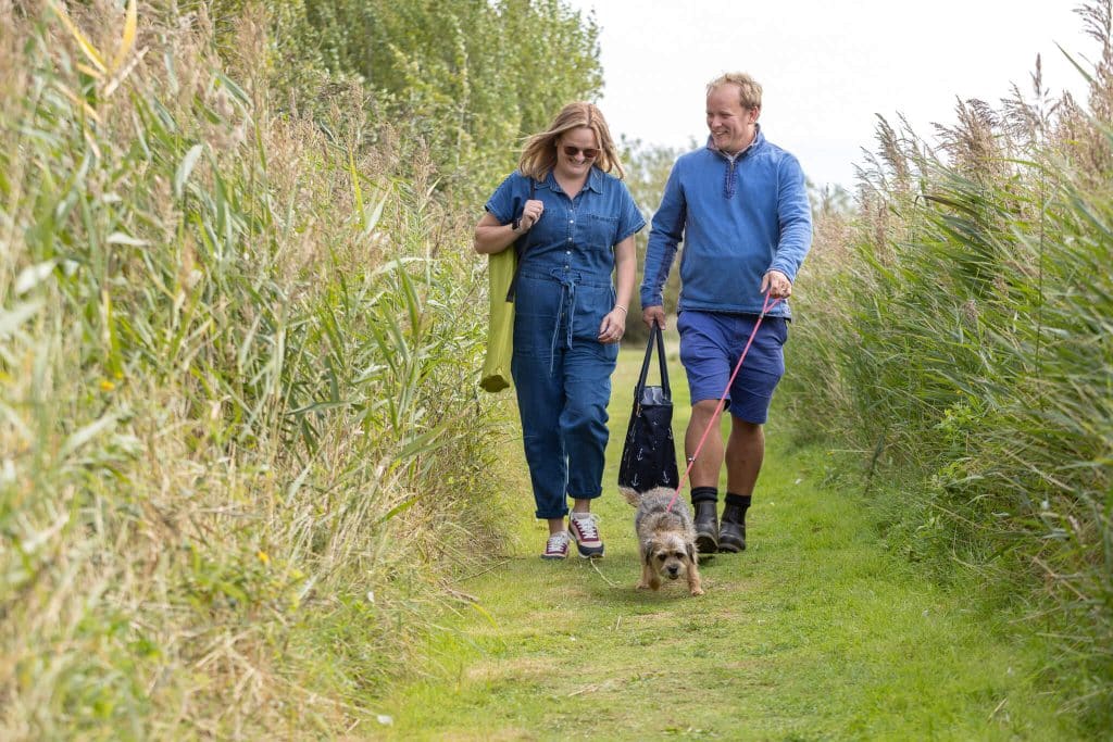 A couple in casual clothing walk, with their dog along a grassy path, smiling.