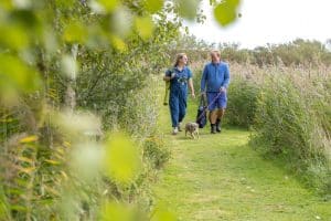 A couple in casual clothing walk, with their dog along a grassy path, smiling.