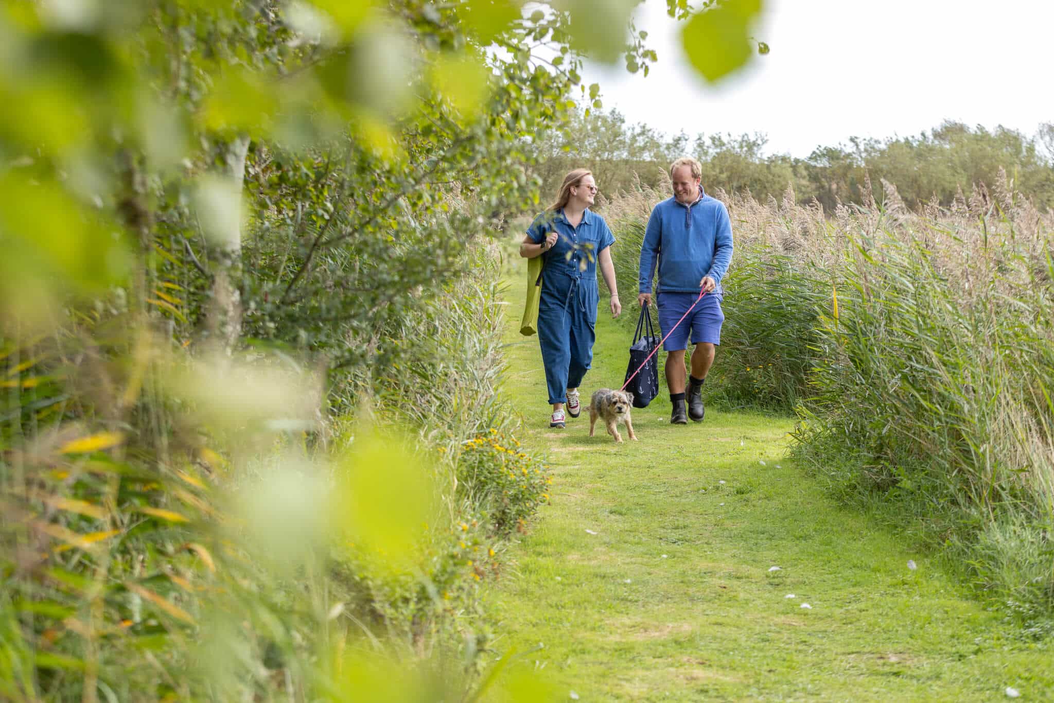 A couple in casual clothing walk, with their dog along a grassy path, smiling.