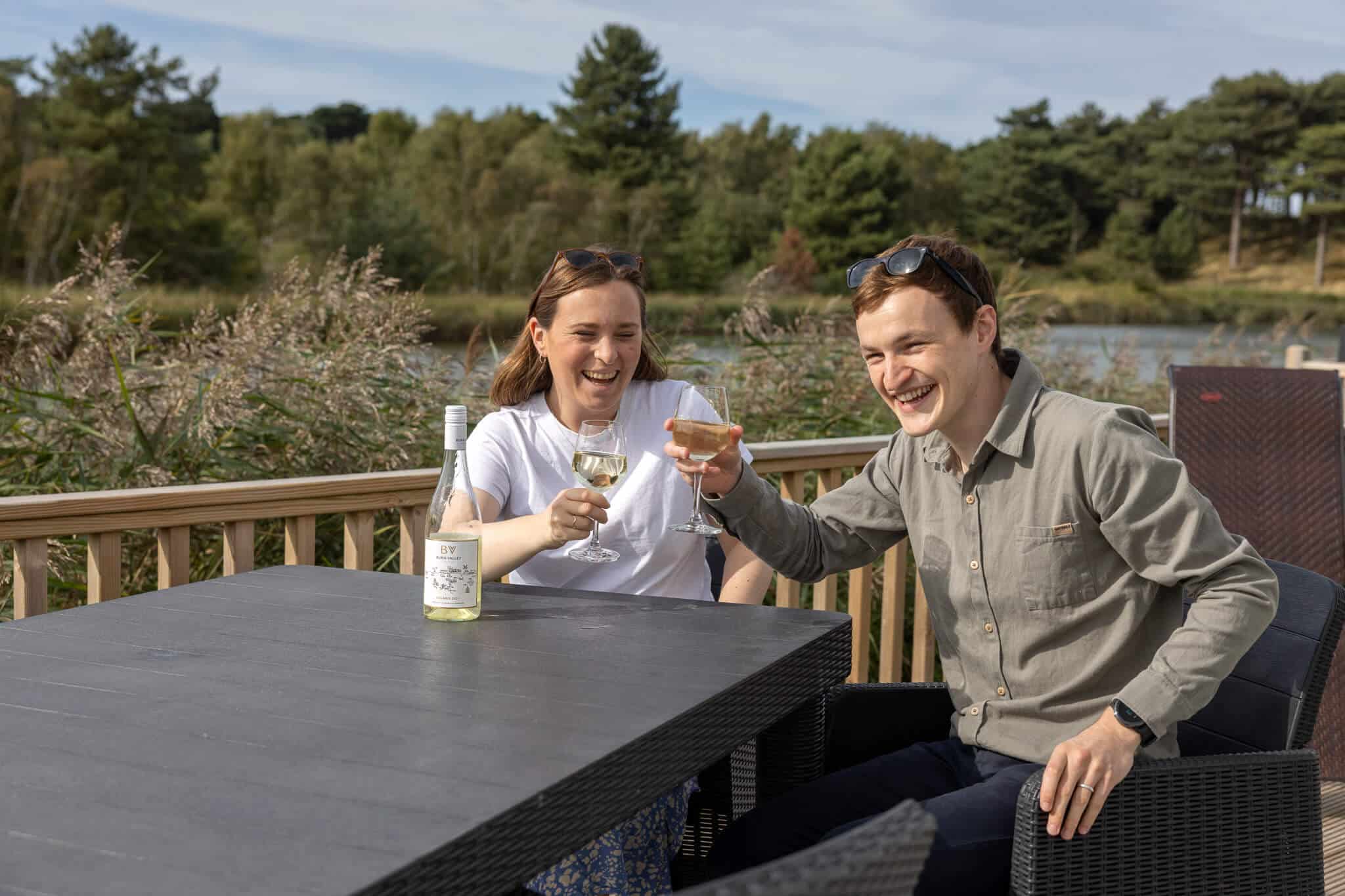 A couple sit at a table and chairs with a glass of white wine each, overlooking a lake. They are laughing.