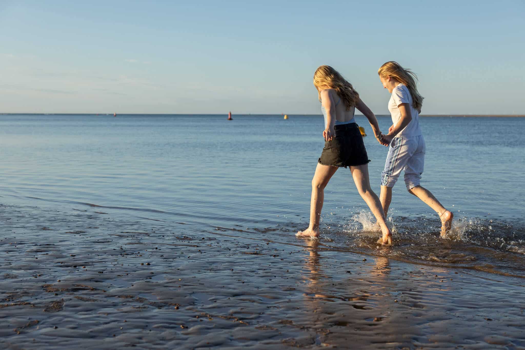 Two girls in summer clothes run in the shallows of the sea.