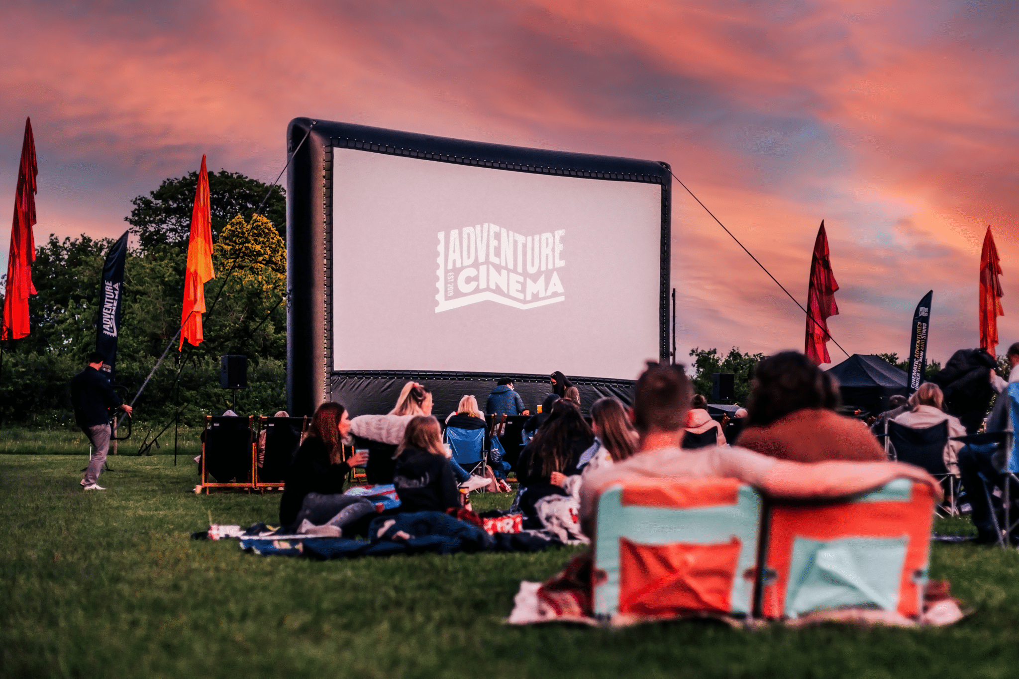 A large outdoor cinema screen against a sunset. People sit on chairs and blankets in front of the screen.