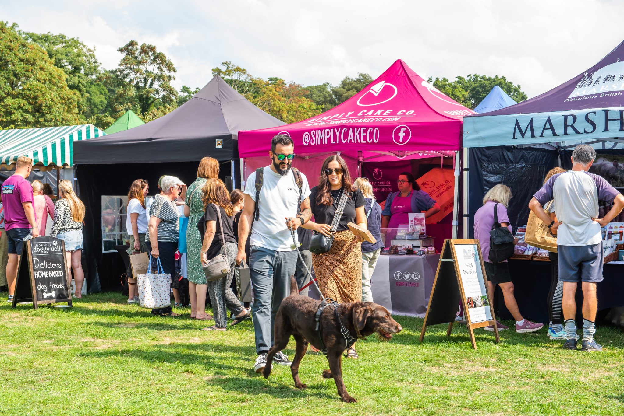 A couple and their dog walk next to a series of food stalls in gazebos on a summer day.