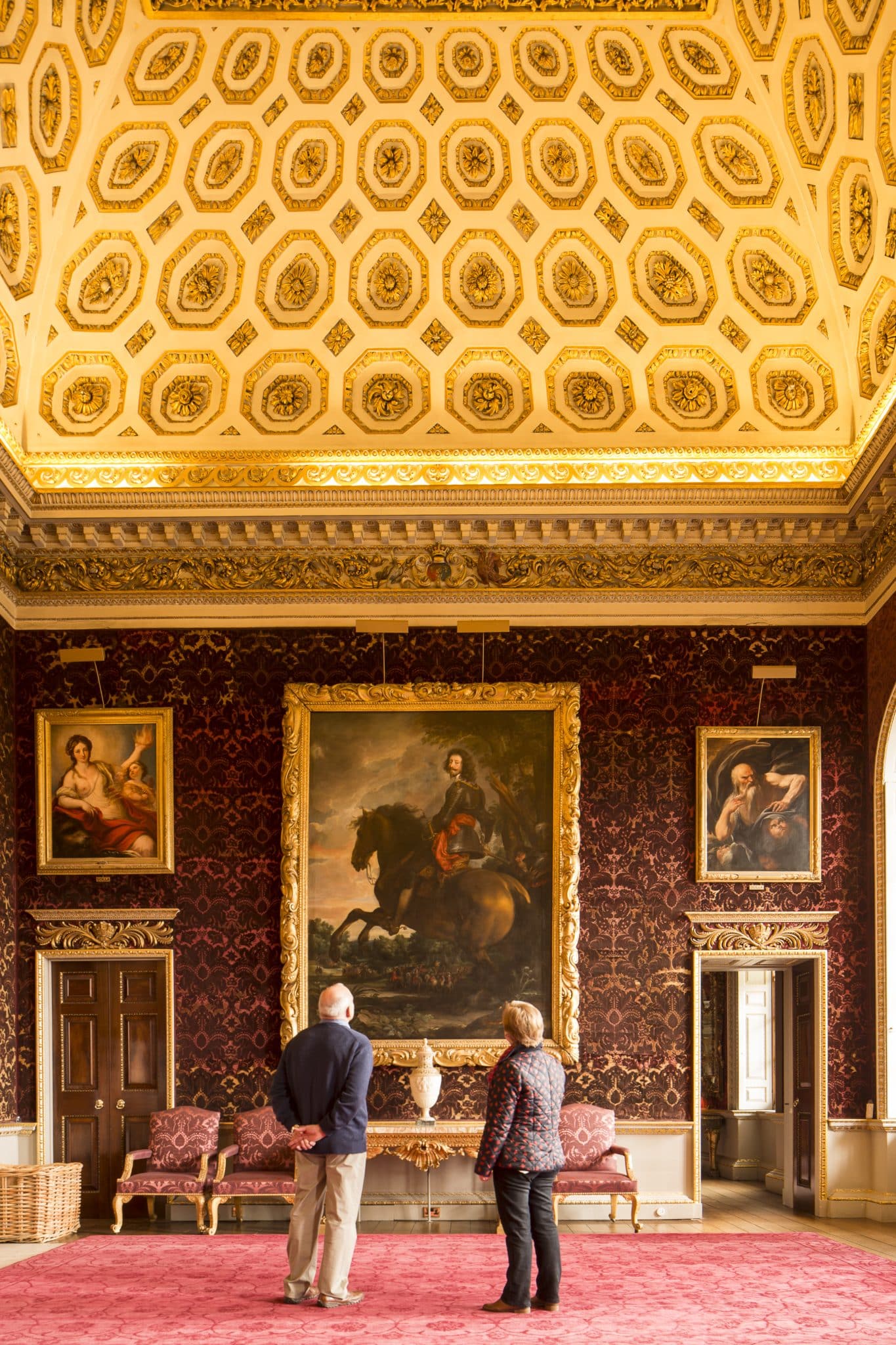 Two people standing in the Saloon at Holkham Hall, looking at a large painting by Van Dyck of the Duke of Arenburg.