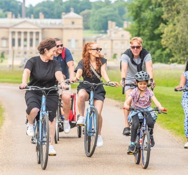 A group of adults and children in summer clothing cycle towards the camera, with Holkham Hall in the background