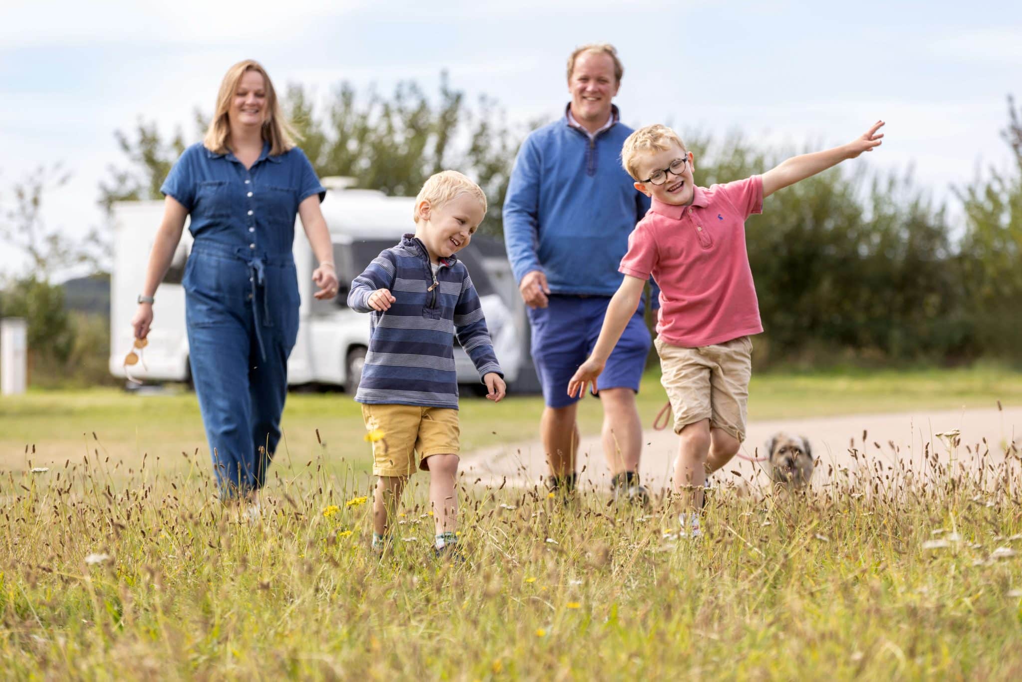 Two young boys and their parents and border terrier walk through the grass at Pinewoods Holiday Park. One boy has his arms out like a plane.