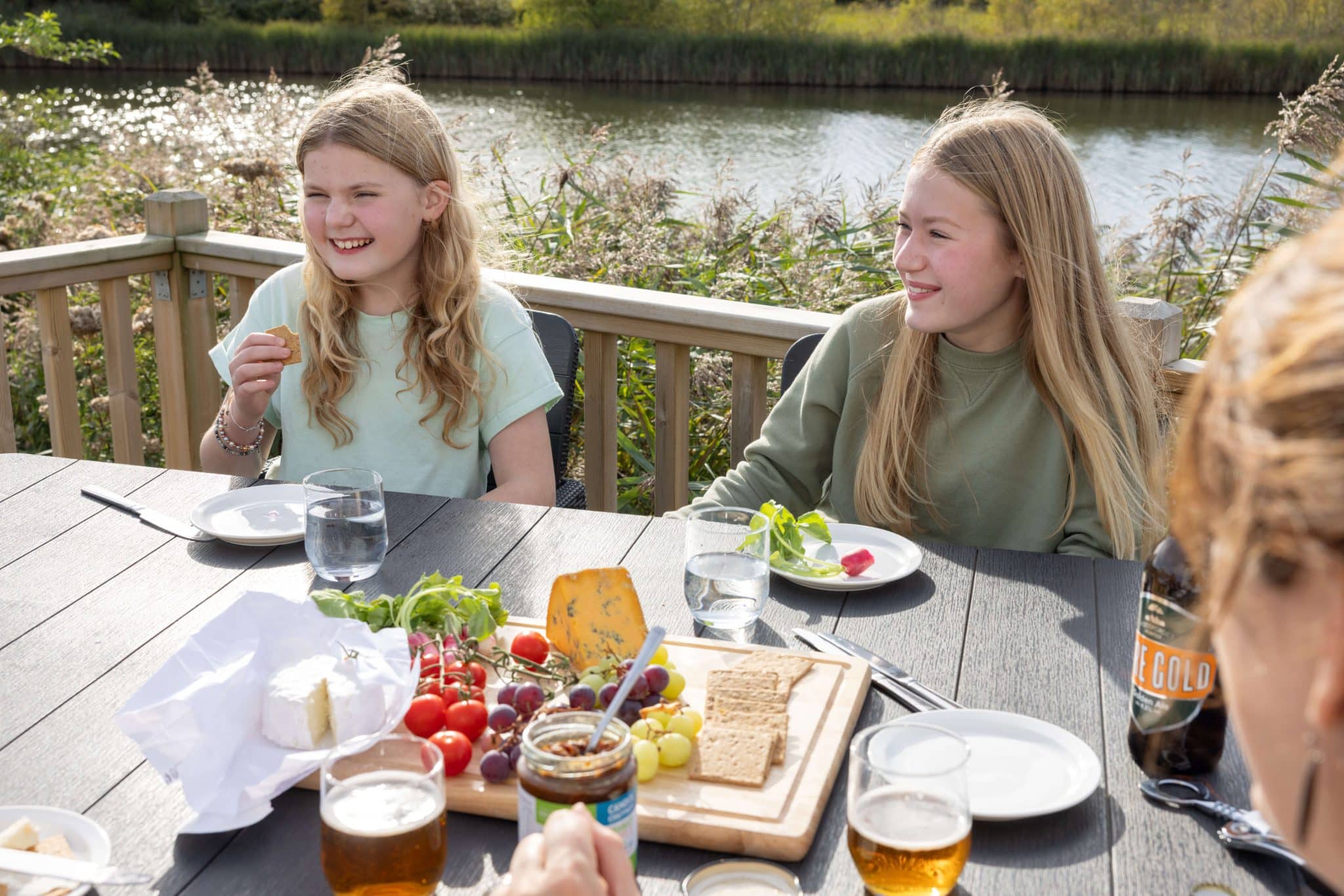 Two teenage girls sit at a table laughing with someone across from them. There is food on the table, and they're sitting outdoors on some decking overlooking a lake.