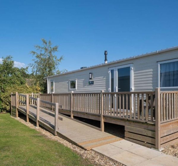 A static caravan next to grass and trees with a wheelchair ramp.