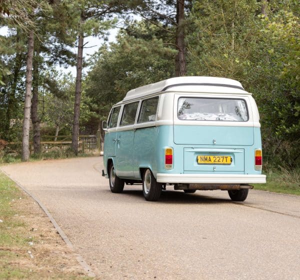 A pale blue, vintage VW campervan drives along a lane through the trees at Pinewoods Holiday Park in Norfolk.