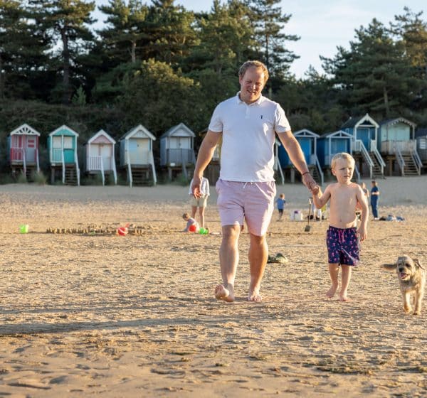 A man and boy in beach clothes walk with their dog on Wells Beach in Norfolk, with colourful beach huts behind them.